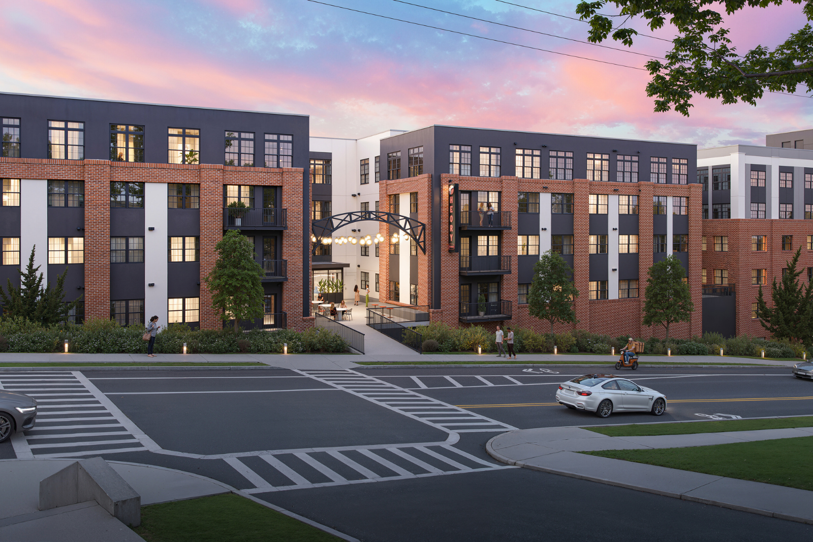 Street-facing view of a modern apartment community with brick-and-panel architecture, illuminated entrance, balconies, landscaped walkways, and pedestrian-friendly streetscape at sunset.