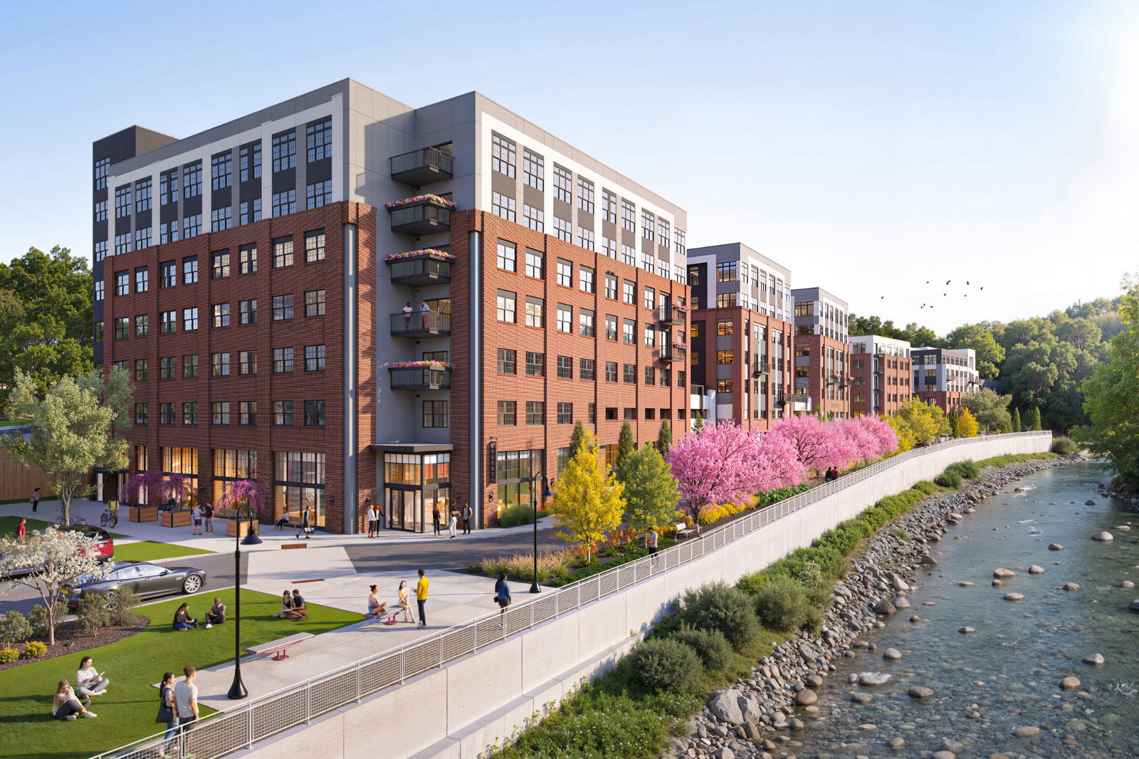 Riverside apartment community featuring contemporary brick buildings, private balconies, landscaped green spaces, walking paths, and scenic waterfront views.
