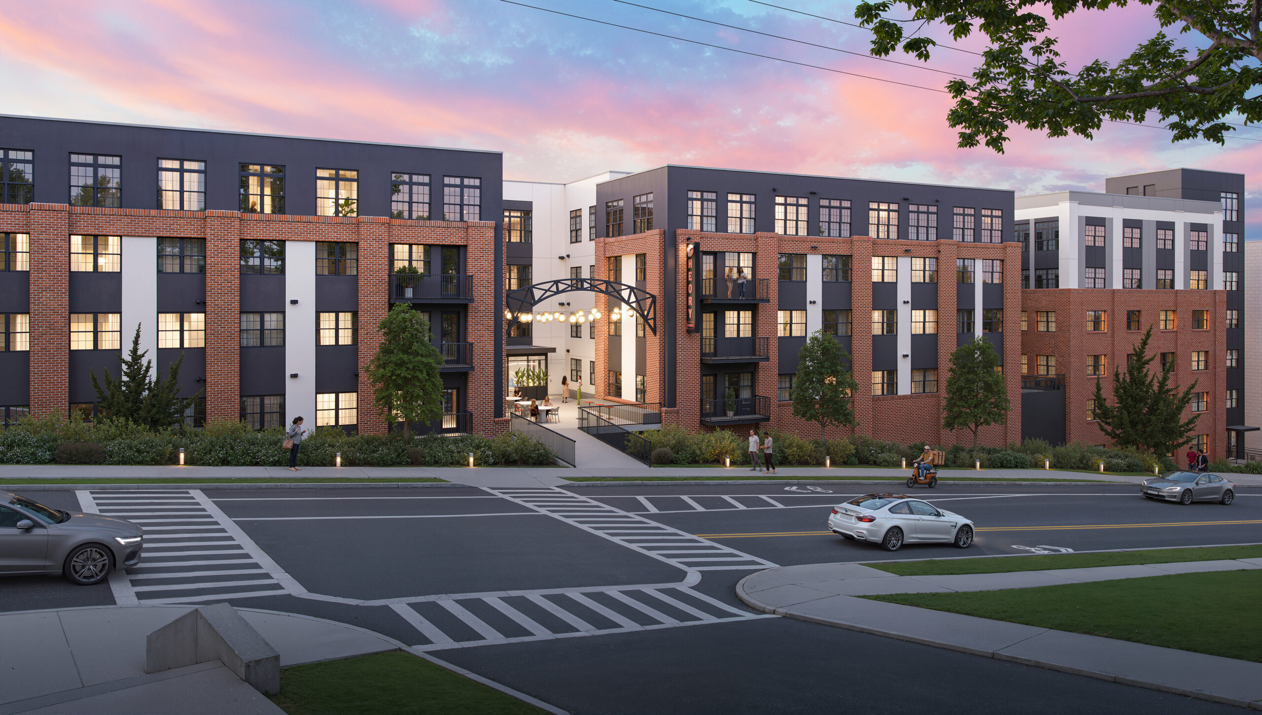 Street-facing view of a modern apartment community with brick-and-panel architecture, illuminated entrance, balconies, landscaped walkways, and pedestrian-friendly streetscape at sunset.